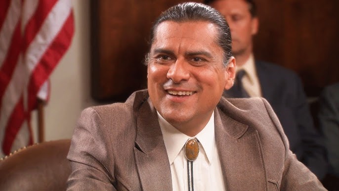 jonathan joss in a brown suit and bolo tie sits at a table in a formal meeting room with people seated behind him.