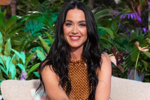 Katy Perry smiling while seated outdoors with greenery in the background.