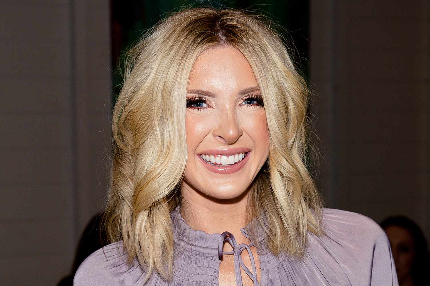 Lindsie Chrisley (now Lindsie Landsman) smiling at a public event, wearing a light purple blouse with wavy blonde hair.