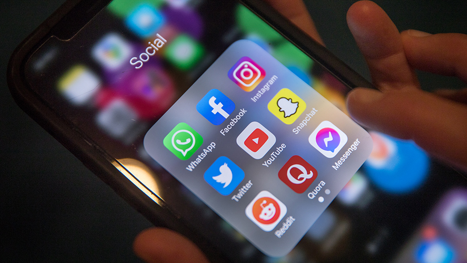 Close-up of a smartphone showing a folder of social media apps, including WhatsApp, Facebook, Instagram, Snapchat, YouTube, Twitter, Reddit and Messenger.