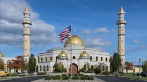 The Islamic Center of America in Dearborn, Michigan, with its gold domes and twin minarets, seen under a clear sky with an American flag in front.