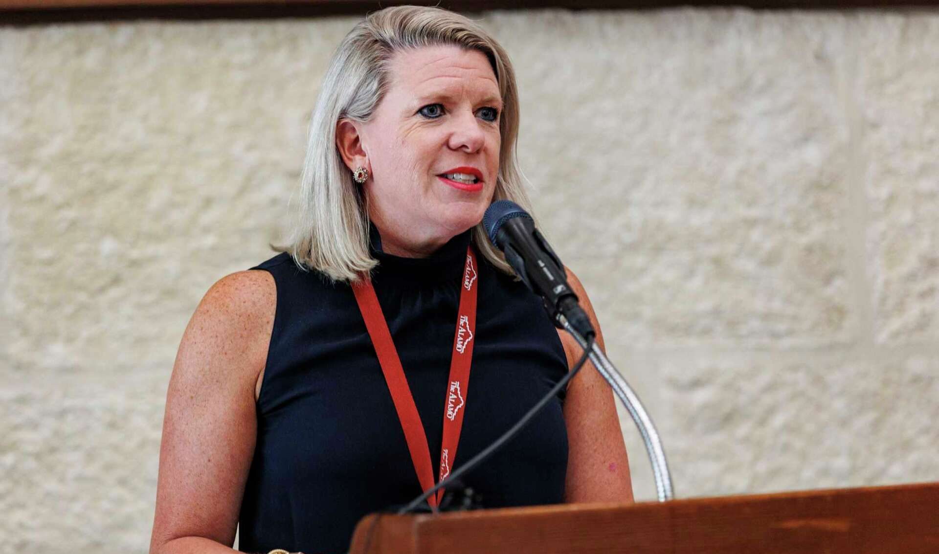 A woman speaks at a podium with a microphone during a public address, wearing a dark sleeveless top and a red lanyard.