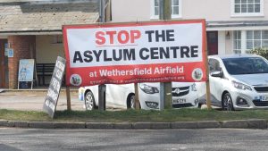 A roadside sign in Wethersfield reading “Stop the Asylum Centre at Wethersfield Airfield,” displayed in front of parked cars and residential buildings.