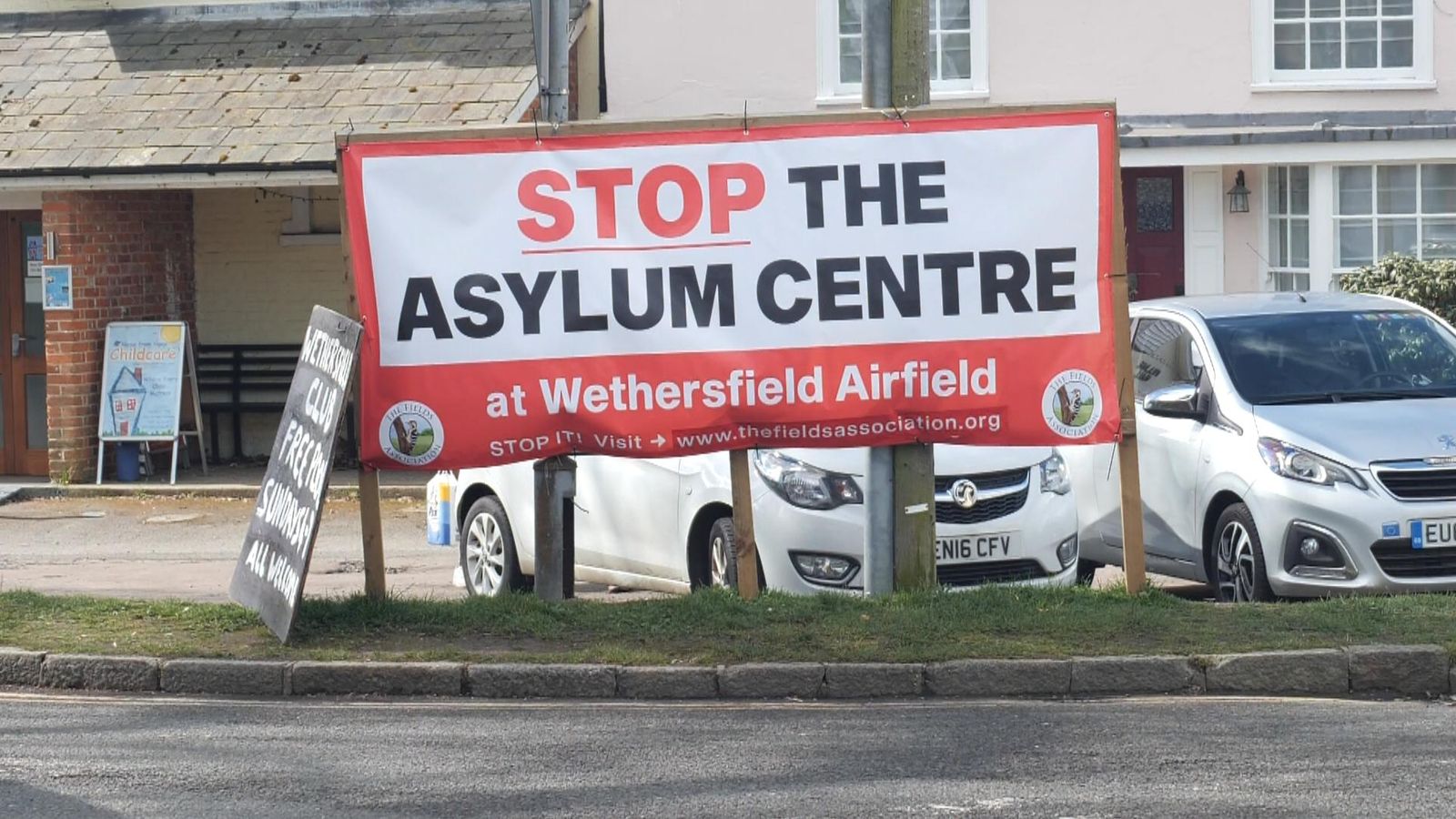 A roadside sign in Wethersfield reading “Stop the Asylum Centre at Wethersfield Airfield,” displayed in front of parked cars and residential buildings.