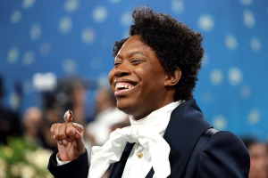 Jeremy O Harris smiling at a public event while wearing a formal navy coat and white necktie.