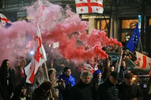 Protesters in Tbilisi march with Georgian and EU flags as red smoke fills the street.