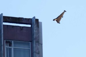 A military drone flies at a low altitude near a damaged high-rise building against a clear sky.