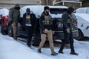 Federal immigration agents wearing tactical gear conduct an enforcement operation in a snowy Minneapolis neighborhood.