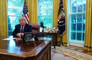 Former U.S. President Donald Trump seated at the Resolute Desk in the Oval Office, with U.S. flags and presidential insignia visible behind him.
